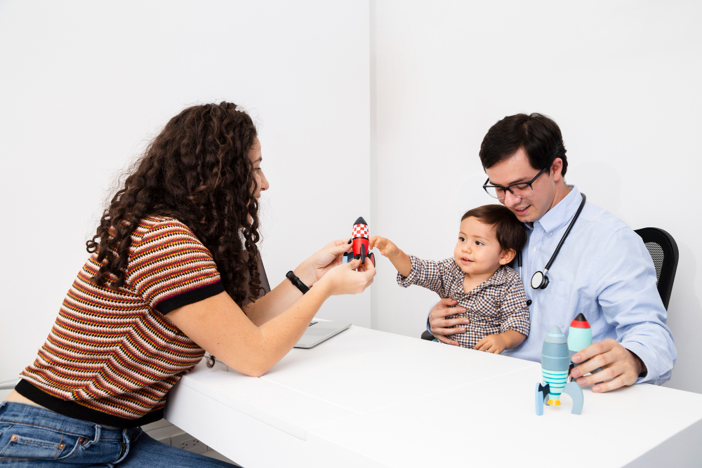 Pediatra atende criança em consulta, enquanto o pai e a mãe interagem com brinquedos sobre a mesa.