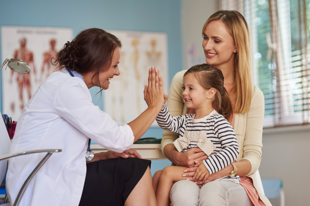 Pediatra cumprimenta criança durante consulta, enquanto a mãe a acompanha sentada no consultório.