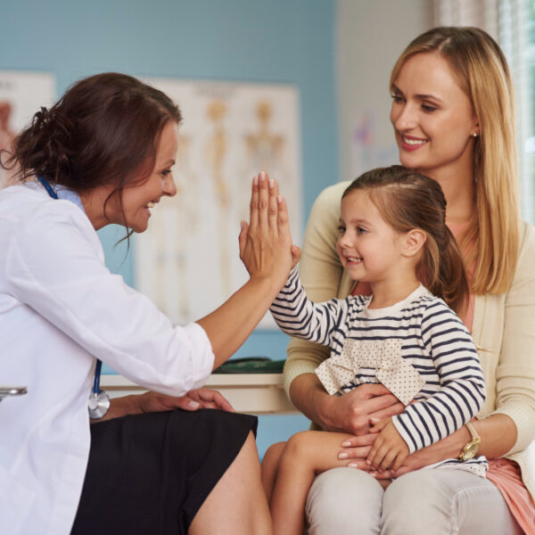 Pediatra cumprimenta criança durante consulta, enquanto a mãe a acompanha sentada no consultório.